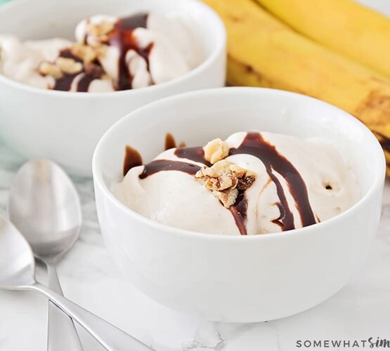 two white bowls filled with frozen banana ice cream topped with chocolate syrup and nuts. On the counter next to the bowls are two spoons and a small bunch of bananas