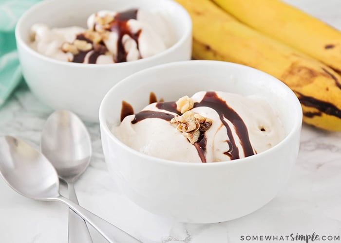 two white bowls filled with frozen banana ice cream topped with chocolate syrup and nuts. On the counter next to the bowls are two spoons and a small bunch of bananas