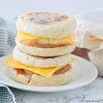 Two make ahead breakfast sandwiches stacked on top of each other on a white plate. Each one is made with an English muffing, American cheese, eggs, and Canadian bacon. Behind the plate are three more sandwiches wrapped in parchment paper.