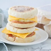 Two make ahead breakfast sandwiches stacked on top of each other on a white plate. Each one is made with an English muffing, American cheese, eggs, and Canadian bacon. Behind the plate are three more sandwiches wrapped in parchment paper.