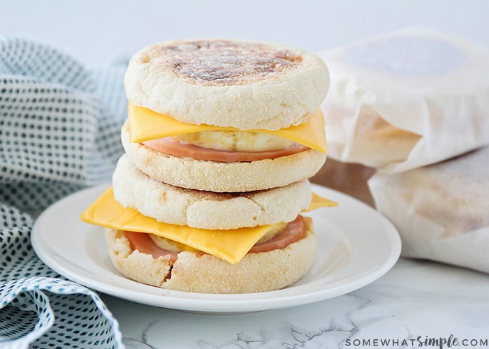 Two make ahead breakfast sandwiches stacked on top of each other on a white plate. Each one is made with an English muffing, American cheese, eggs, and Canadian bacon. Behind the plate are three more sandwiches wrapped in parchment paper.