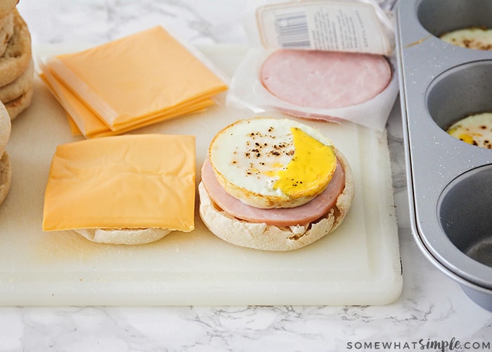 a breakfast sandwich being assembled on a cutting board. On one half of the English muffin is a slice of American cheese. On the other half is an egg and a slice of Canadian bacon. 