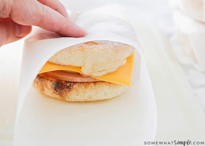 a hand wrapping a breakfast sandwich with parchment paper. The sandwich has a slice of American cheese, ham and eggs.