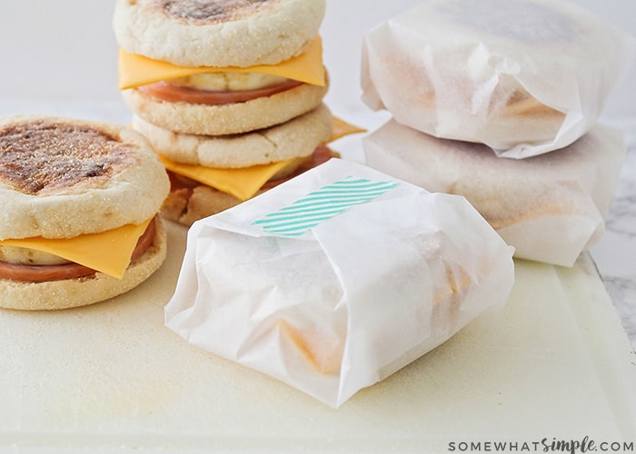 multiple breakfast sandwiches are stacked on a cutting board and half are wrapped in parchment paper.