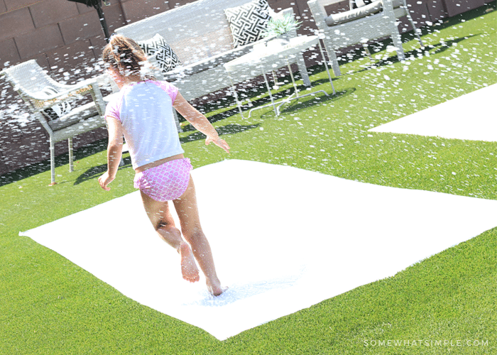 a little girl running through spraying water on a diy slip and slide made from a plastic drop cloth