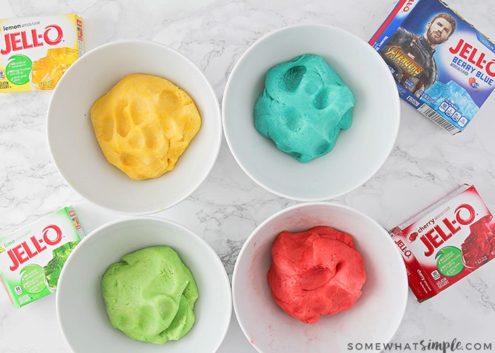 looking down on 4 bowls of jello cookie dough. Each bowl has one color dough, yellow, blue, green and red and on the counter next to them is a box of jello that was used for each color