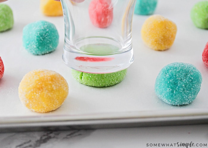 balls of bright colorful cookie dough on a baking sheet before they go in the oven. A glass is being pressed down on one of the cookie dough balls to flatten them before baking.