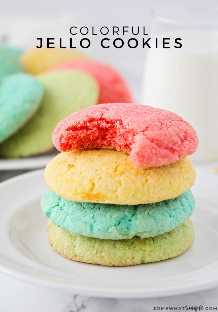 a stack of bright red, yellow, blue and green jello cookies on a white plate with a jar of milk and a plate of more cookies in the background with the words colorful jello cookies overlayed at the top of the picture