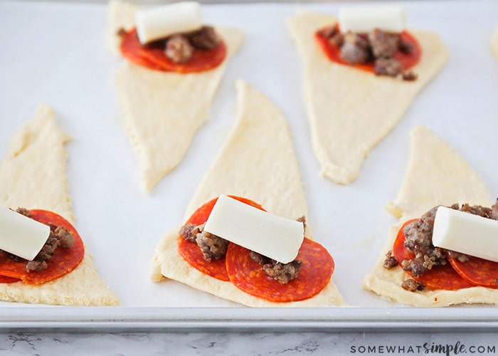 multiple precooked pizza bites on a cookie sheet with slices of pepperoni, cheese and sausage on raw dough
