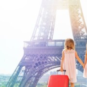 two young girls in pick dresses standing in front of the Eiffel Tower. The younger one has a red suitcase filled items from a packing list for europe.