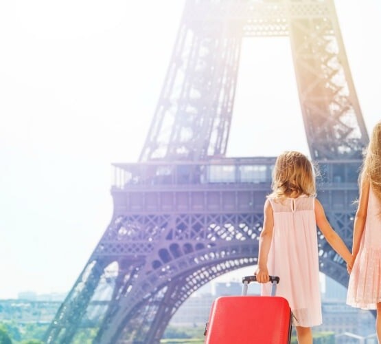 two young girls in pick dresses standing in front of the Eiffel Tower. The younger one has a red suitcase filled items from a packing list for europe.