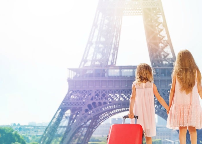 two young girls in pick dresses standing in front of the Eiffel Tower. The younger one has a red suitcase filled items from a packing list for europe.
