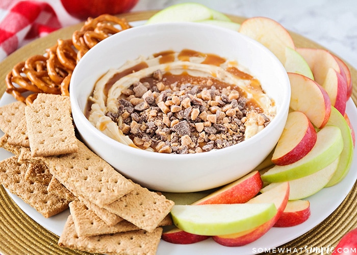 a bowl full of caramel apple dip with bits of candy bar on top with apple slice, graham crackers and pretzels around the bowl on a serving tray