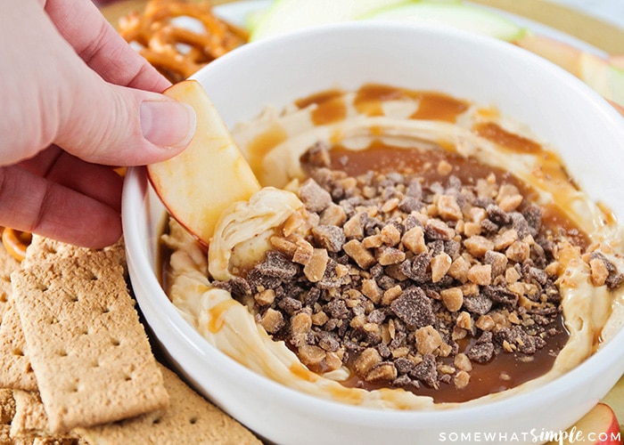 a hand dipping an apple slice into this caramel cream cheese dip topped with Heath bar bits