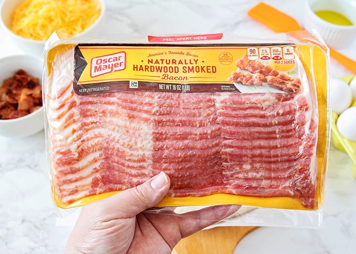 a hand holding a package of oscar mayer bacon is one of the main ingredients for this breakfast pizza