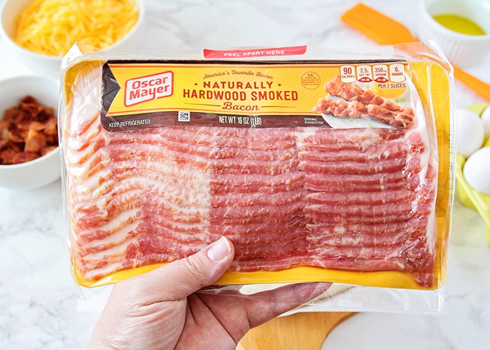 a hand holding a package of oscar mayer bacon is one of the main ingredients for this breakfast pizza