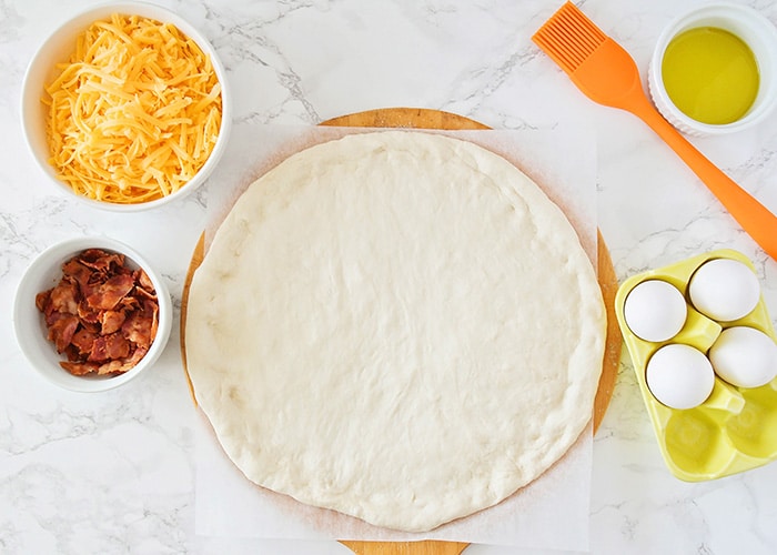 looking down on the ingredients to make breakfast pizza; cheese, bacon, pizza dough, oil and eggs