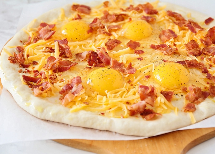 a bacon breakfast pizza topped with bacon, cheese and raw eggs before it is cooked in the oven
