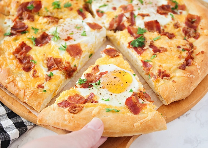 a hand pulling out a slice from a breakfast pizza that's topped with bacon, eggs, cheese and cilantro