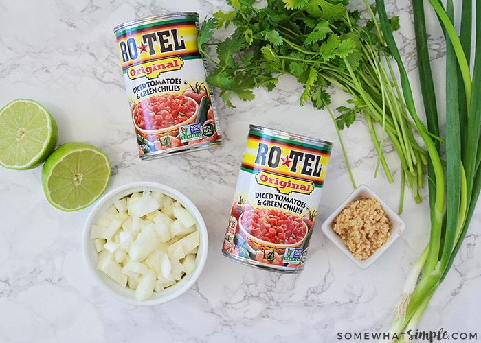 two cans of Ro Tel diced tomatoes with green chilies, a lime cut in half, a bowl of diced onions, a stalk of cilantro, a bowl of minced garlic and a stalk of green onions laying on the counter which are the ingredients used to make this recipe.