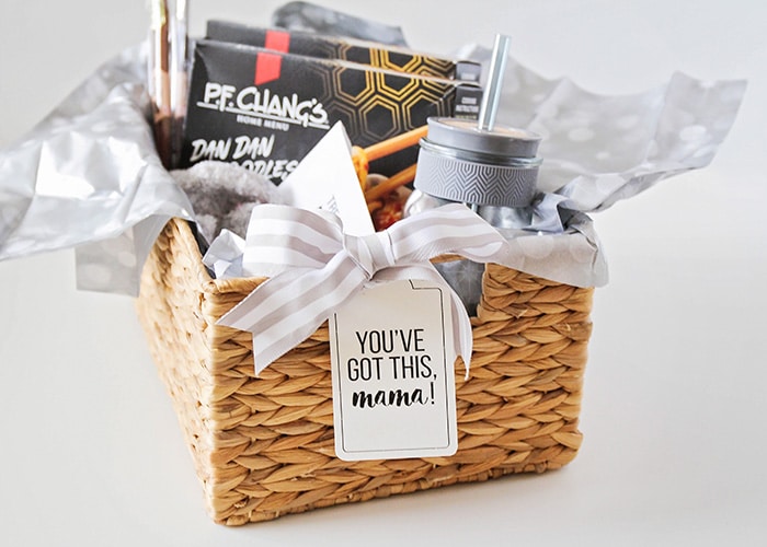 a new mom gift basket filled with gift items wrapped in gray tissue paper and a light grey striped bow with a tag on the front that says you've got this mama!