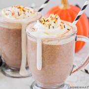 two glass mugs filled with a homemade pumpkin spice hot chocolate topped with whipped cream and sprinkles
