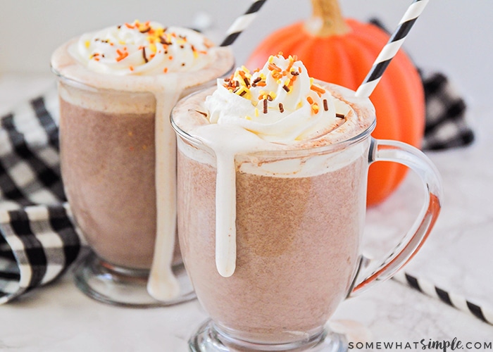 two glass mugs filled with a homemade pumpkin spice hot chocolate topped with whipped cream and sprinkles