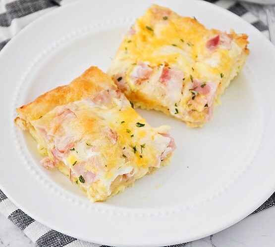 two squares of a croissant breakfast egg casserole made with ham on a white plate sitting on top of a cloth black and white striped napkin.