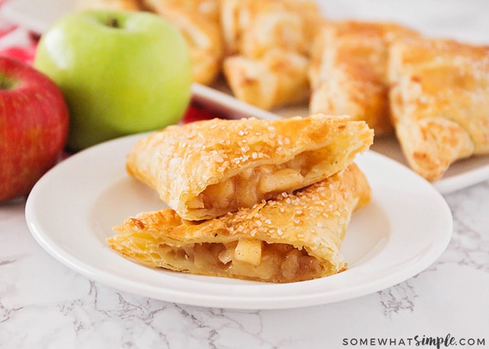 two apple turnovers with a sweet cinnamon apple filling encased in flaky puff pastry on a white plate with a red and green apple with additional turnovers in the background
