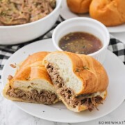 a french dip sandwich cut in half and stacked on a white plate with a side of au jus in a small white bowl. Behind the plate is a bowl filled with shredded beef and a plate with French rolls on it.