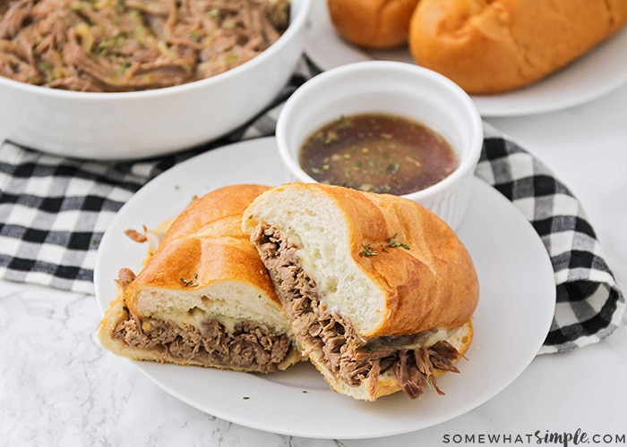 a french dip sandwich cut in half and stacked on a white plate with a side of au jus in a small white bowl. Behind the plate is a bowl filled with shredded beef and a plate with French rolls on it.