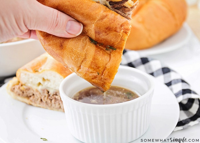 a hand dipping a half of a crock pot French dip sandwich into a small white bowl of au jus with the other half of the sandwich in the background.