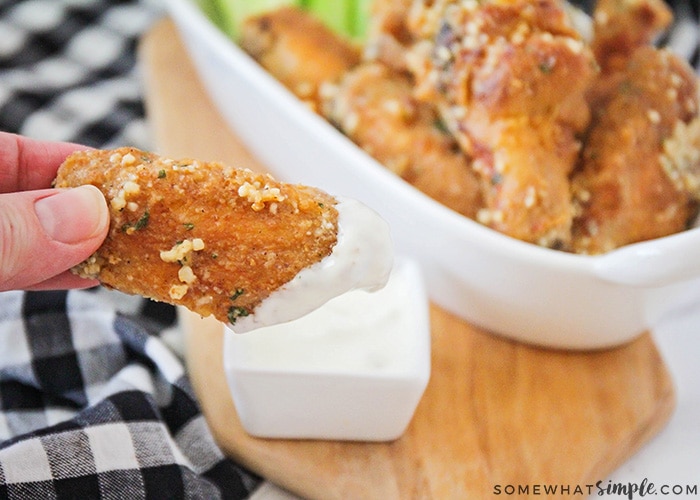 a chicken wing backed in garlic and parmesan being dipped in a side of ranch dressing. A bowl of several chicken wings are in the background.