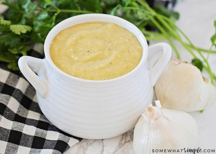 a white gravy boat filled with homemade green enchilada sauce. Next to the gravy boat are two white onions, a parsley bunch and a black and white checkered cloth napkin.