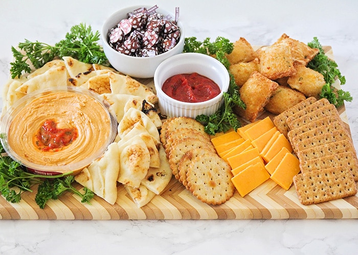 An easy to assemble kid friendly appetizer board that is filled with slices of flatbread, various crackers, slices of cheddar cheese, baked pizza bites surrounded by parsley with a white dish filled with Hershey's kisses, another white dish in the middle filled with marinara sauce and a small glass bowl filled with hummus.