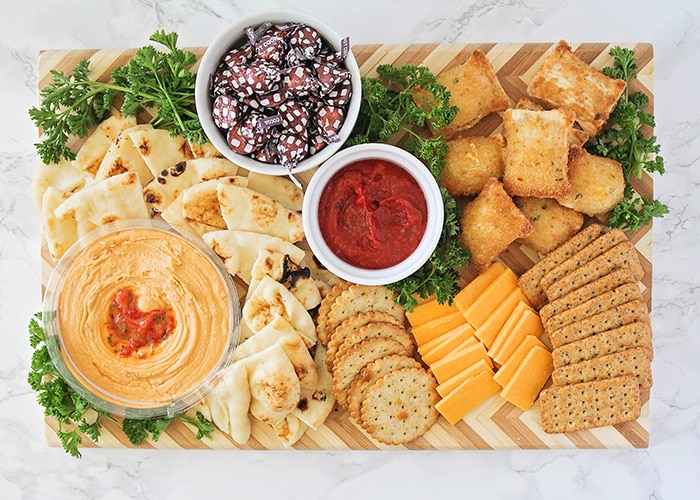 looking down on a board filled with the best appetizers for kids. Included on the board are slices of flatbread, various crackers, slices of cheddar cheese, baked pizza bites surrounded by parsley with a white dish filled with Hershey's kisses, another white dish in the middle filled with marinara sauce and a small glass bowl filled with hummus.
