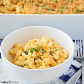 a white bowl filled with baked macaroni and cheese topped with bread crumbs and parsley. Behind the bowl is a casserole pan filled with more macaroni and cheese. Next to the bowl are two forks in a crossed patter laying on top of a blue and white checkered cloth napkin.