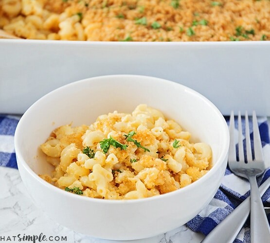 a white bowl filled with baked macaroni and cheese topped with bread crumbs and parsley. Behind the bowl is a casserole pan filled with more macaroni and cheese. Next to the bowl are two forks in a crossed patter laying on top of a blue and white checkered cloth napkin.