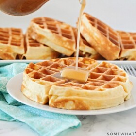 a Belgian waffle on a white plate topped with a pad of butter. Maple syrup is being poured from above onto the waffle. Behind the plate on the counter is a long serving tray filled with additional golden brown Belgian waffles.