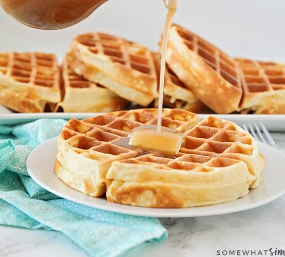 a Belgian waffle on a white plate topped with a pad of butter. Maple syrup is being poured from above onto the waffle. Behind the plate on the counter is a long serving tray filled with additional golden brown Belgian waffles.