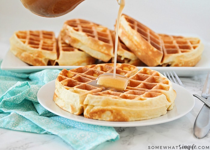 a Belgian waffle on a white plate topped with a pad of butter. Maple syrup is being poured from above onto the waffle. Behind the plate on the counter is a long serving tray filled with additional golden brown Belgian waffles.