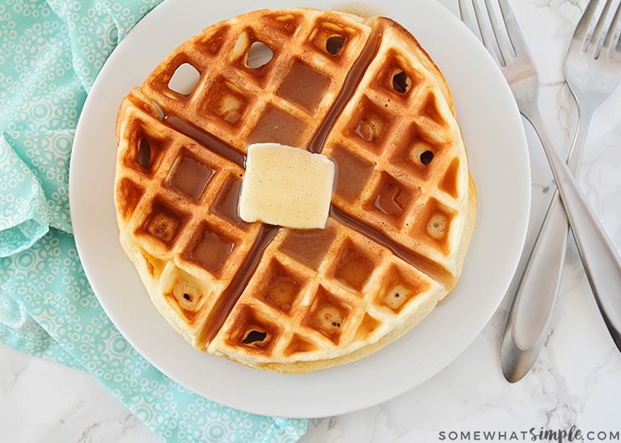 looking down on a plate with a homemade Belgian waffle topped with a pad of butter and maple syrup. Next to the plate are two forks crossed on top of each other.