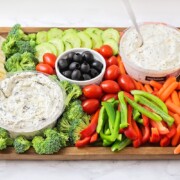 a colorful veggie tray idea that's filled with crackers, cheese, broccoli pieces, cucumber slices, cherry tomatoes, olives, red and green bell pepper slices, baby carrots and two vegetable dips all on a dark acacia wood serving tray.