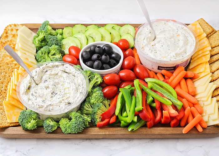 a colorful veggie tray idea that's filled with crackers, cheese, broccoli pieces, cucumber slices, cherry tomatoes, olives, red and green bell pepper slices, baby carrots and two vegetable dips all on a dark acacia wood serving tray. 