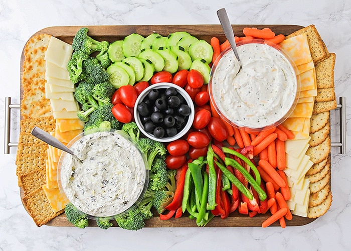 looking down on a simple and colorful veggie party tray idea that's filled with crackers, cheese, broccoli pieces, cucumber slices, cherry tomatoes, olives, red and green bell pepper slices, baby carrots and two vegetable party tray dips all on a dark acacia wood serving tray. 