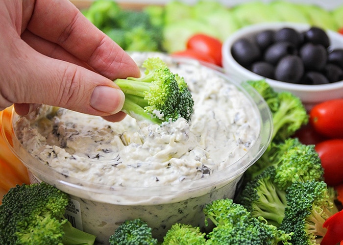 a hand dipping a piece of broccoli into a veggie tray dip. More pieces of broccoli surround the vegetable dip as well as cherry tomatoes and cheese with a small bowl of black olives and cucumber slices in the background on the tray.