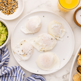 white plate with 5 Poached Eggs on it, surrounded by other breakfast foods