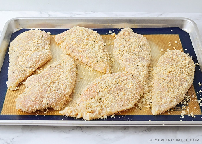 six uncooked chicken breast on a baking sheet covered in panko and bread crumbs