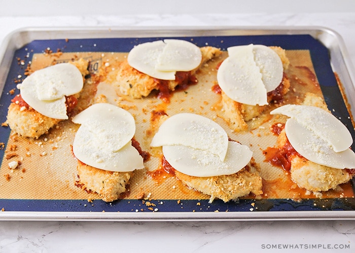 six baked chicken breasts on a baking sheet topped with marinara sauce and slices of provolone cheese placed on top