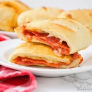 a homemade pepperoni pizza pocket that has been cut in half and the two halves are stacked on top of each other. In the background is a tray filled with several more golden brown pizza pockets.